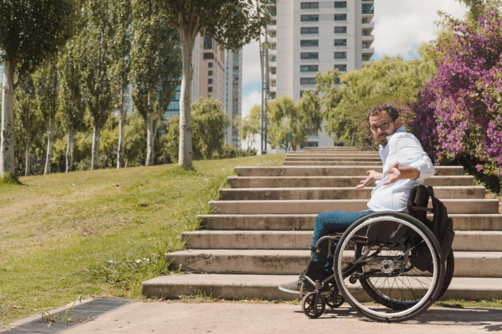Homem usando cadeira de rodas indignado em frente a uma escadaria sem acesso com rampa, representando a importância das cidades inclusivas