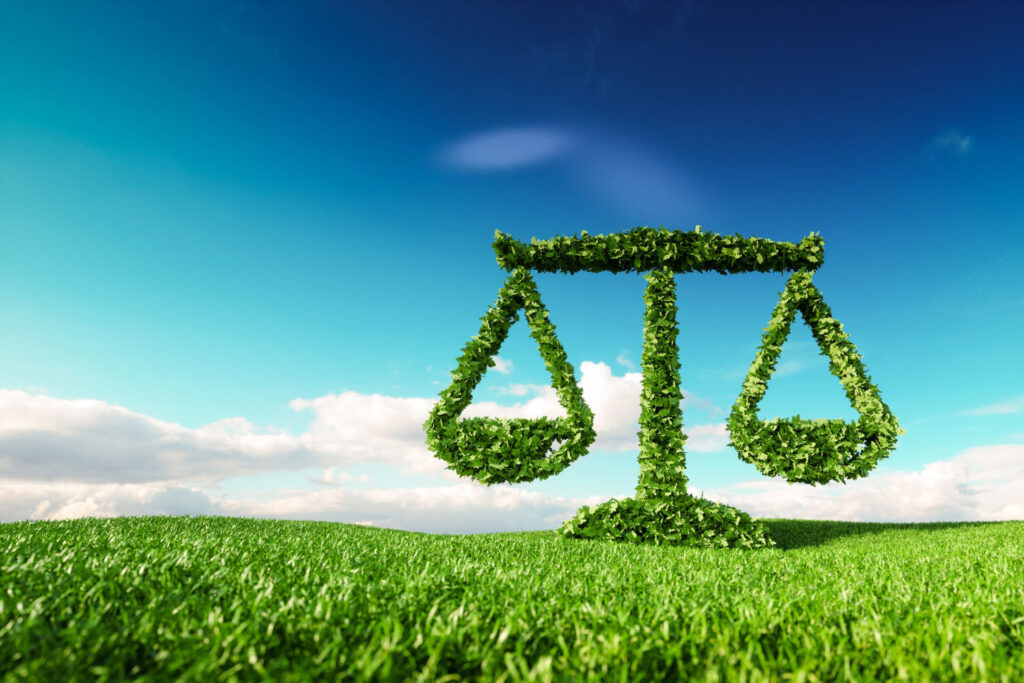 Balança da justiça revestida de folhas verdes sobre um gramado e com céu azul de fundo representando as legislações ambientais.